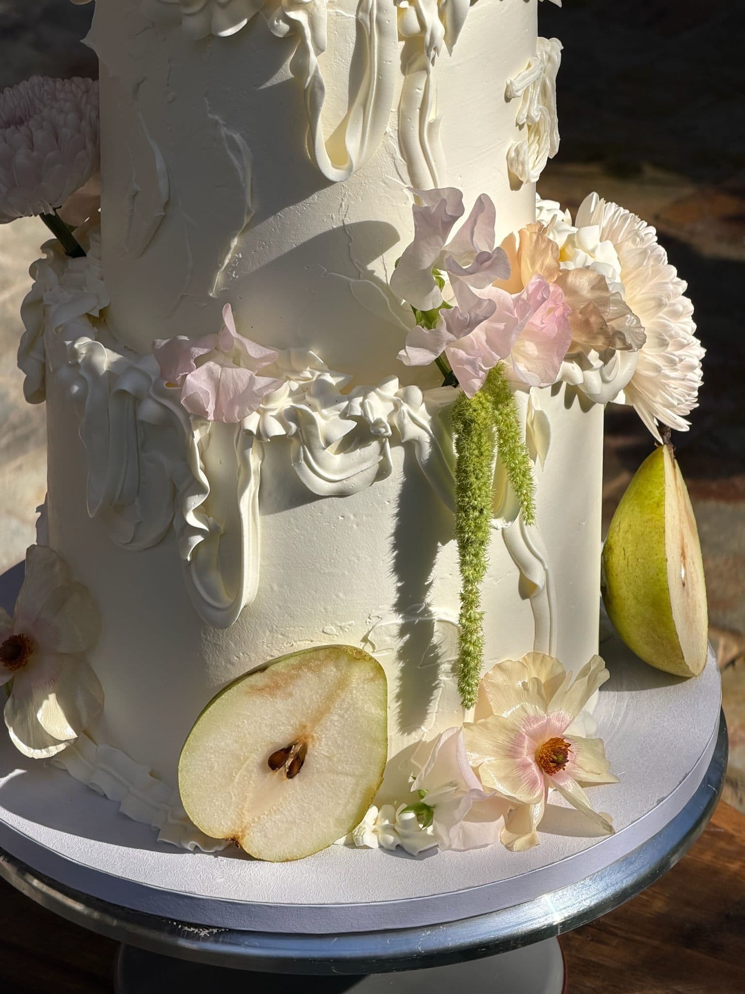 Two-tier cake with sculpted buttercream drapes, sliced pears and blush chrysanthemums, in low sun