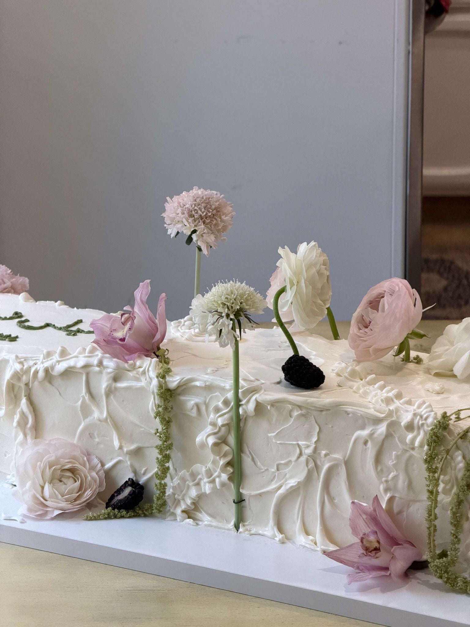 Long ivory sheet cake with pale roses, scabiosa and blackberries set into the buttercream, beside a mirror