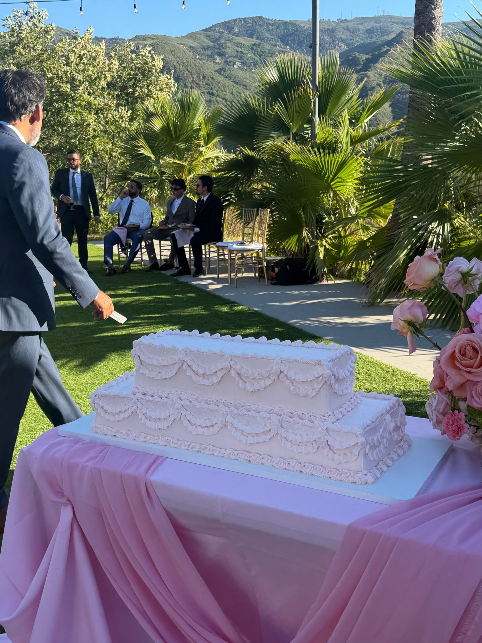Two-tier blush square wedding cake with piped scalloped swags, on a draped table outdoors