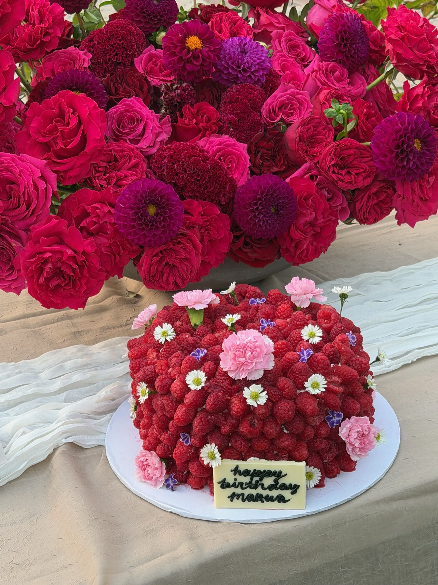 Cake blanketed edge to edge in fresh raspberries, dotted with carnations and daisies, against magenta roses and dahlias