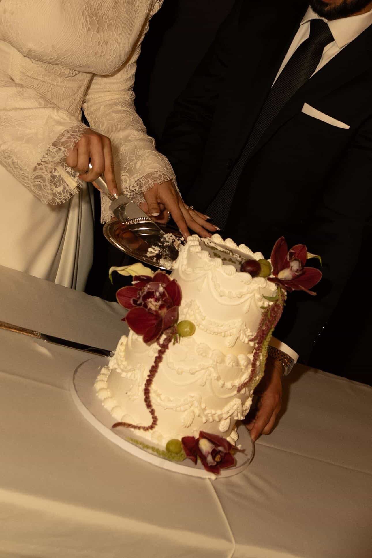 Two-tier wedding cake with piped swags, deep red orchids and grapes, being cut by the couple